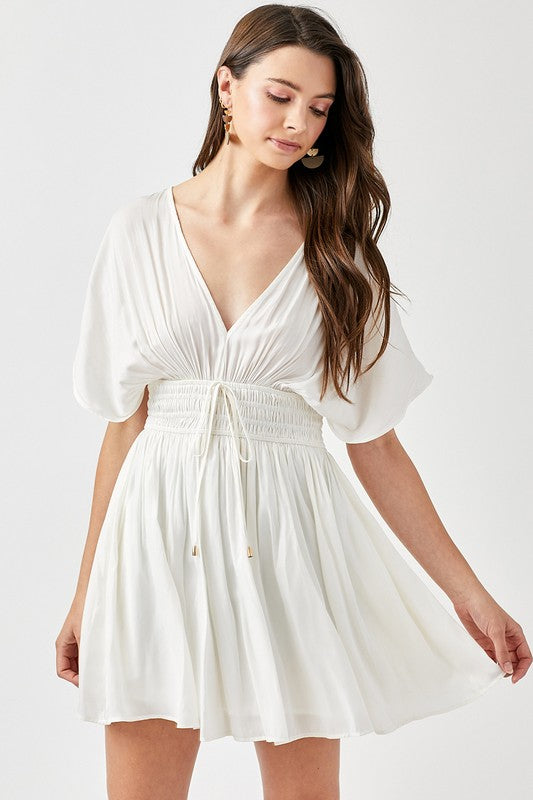 A woman is wearing a Smocked Waist with Tassel Strap Dress, featuring a white color, short sleeves, and a V-neckline, as she stands against a plain background.