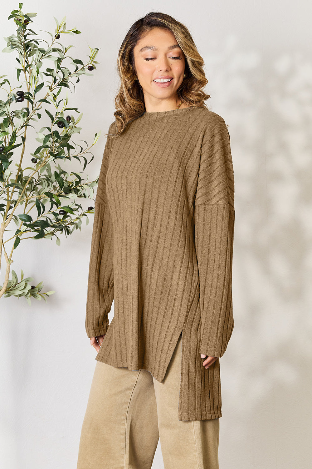 A person with shoulder-length hair smiles while wearing a beige ribbed sweater called the Basic Bae Full Size Ribbed Round Neck Long Sleeve Slit Top, along with matching beige pants, showcasing a basic style. They stand next to a green plant.