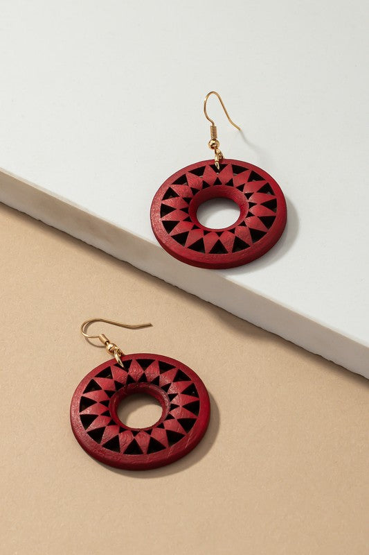 The NEW COLORS Cutout wood circle drop earrings, showcasing exquisite geometric patterns in black, brown, yellow, and orange, are elegantly arranged on a white surface.