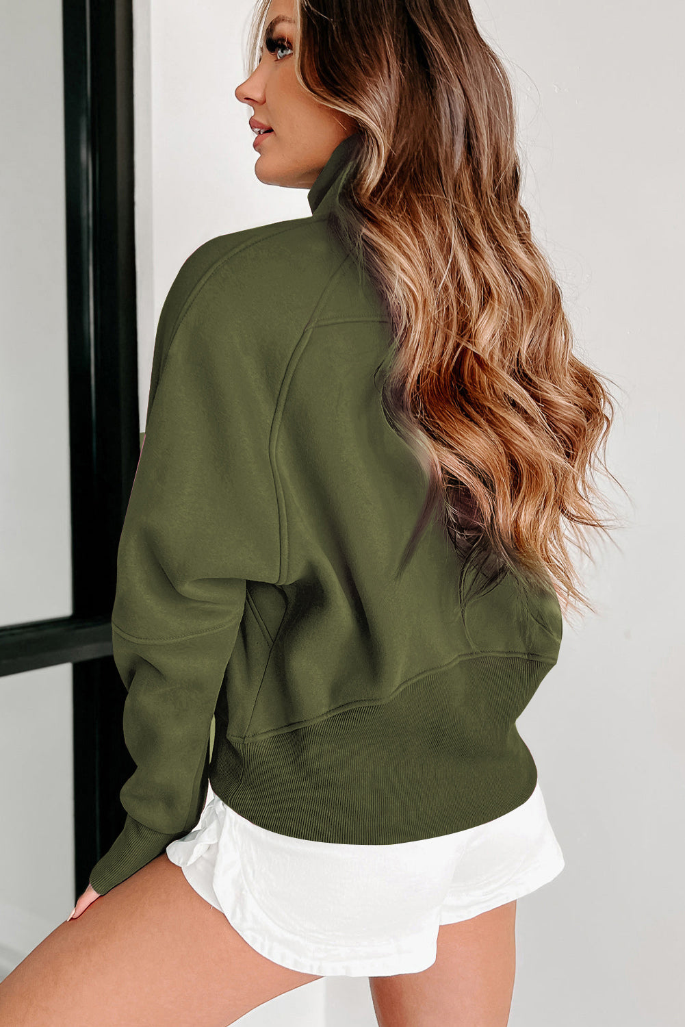 A person with long, wavy hair stands in front of a white and black background, wearing the Green Fleece Lined Zip Up Stand Collar Thumbhole Sleeve Sweatshirt paired with white shorts, creating a stylish look perfect for casual outings.