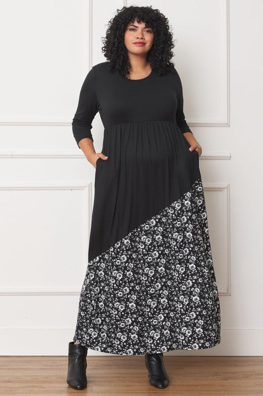 A woman with curly hair is wearing the Asymmetrical Leopard Accent Maxi Dress, featuring a long black design with a floral patterned section at the bottom. She stands against a white paneled wall, her hands tucked in her dress pockets, showcasing its elegant design.