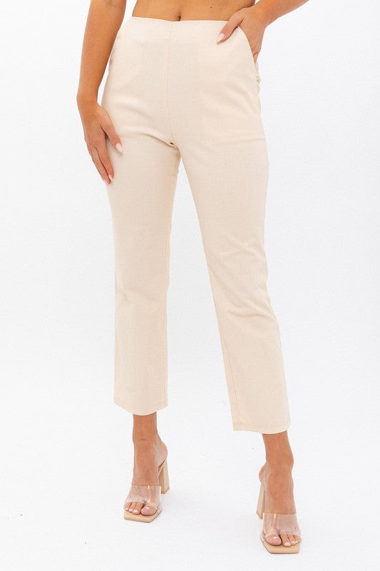 Person wearing High-Waisted Crop Pants in beige with a cropped length and clear high-heeled sandals stands against a white background. One hand is on their hip.