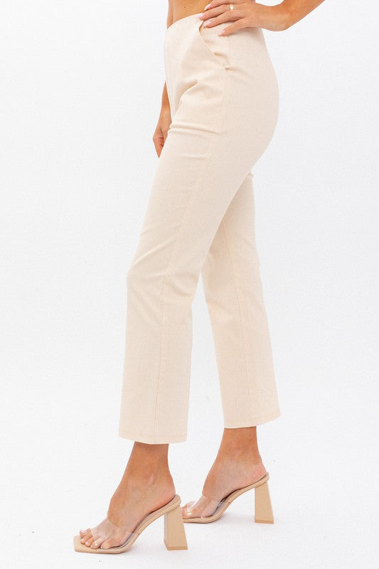 Person wearing High-Waisted Crop Pants in beige with a cropped length and clear high-heeled sandals stands against a white background. One hand is on their hip.