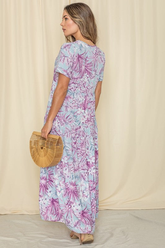 A woman in a Tropical Tiered Flowy Maxi Dress holds a woven handbag, standing against a beige backdrop with a neutral expression.