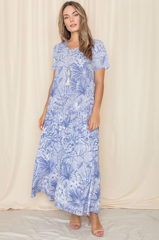 A woman stands against a beige backdrop, showcasing the Tropical Tiered Flowy Maxi Dress, which features a light blue floral polyester design.