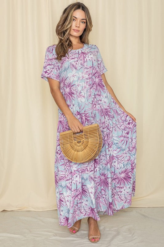 A woman in a Tropical Tiered Flowy Maxi Dress holds a woven handbag, standing against a beige backdrop with a neutral expression.