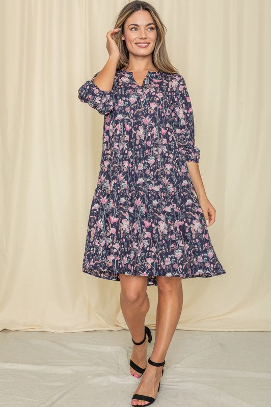 A person elegantly poses in front of a light curtain backdrop, wearing the Floral Notch Neck Midi Dress, crafted from 100% polyester and featuring three-quarter sleeves.