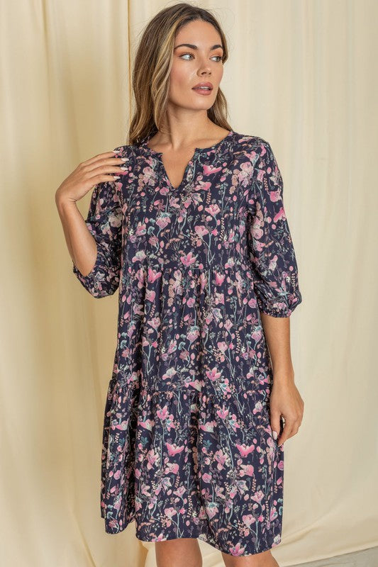 A person elegantly poses in front of a light curtain backdrop, wearing the Floral Notch Neck Midi Dress, crafted from 100% polyester and featuring three-quarter sleeves.