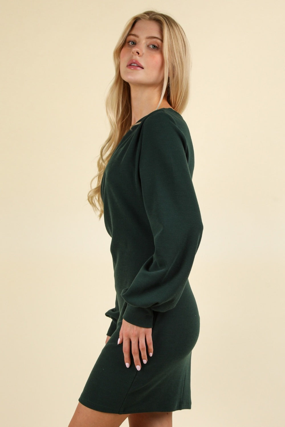 A person stands facing the camera, wearing the VERY J Ruched Detail Bodycon Mini Dress in dark green, featuring a V-neck and a wrap-style bodice. The figure-hugging silhouette accentuates their form beautifully, set against a plain, light-colored background.