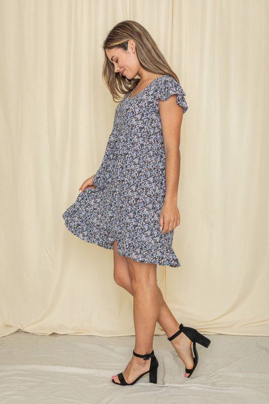 A woman stands in front of a light curtain, wearing the Mix Floral Flutter Sleeve Mini Dress and black heels, smiling slightly.