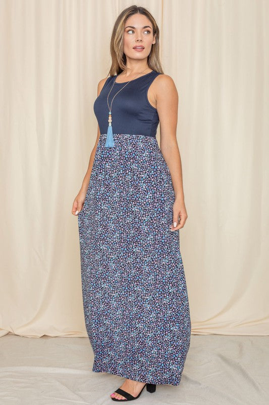 A woman elegantly stands in front of a draped curtain, wearing the Plus Sleeveless Floral Maxi Dress, which features a vibrant red top paired with a flowing blue skirt.