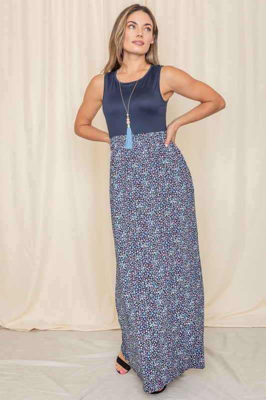 A woman elegantly stands in front of a draped curtain, wearing the Plus Sleeveless Floral Maxi Dress, which features a vibrant red top paired with a flowing blue skirt.