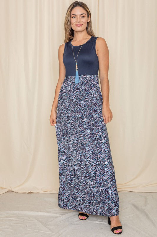 A woman elegantly stands in front of a draped curtain, wearing the Plus Sleeveless Floral Maxi Dress, which features a vibrant red top paired with a flowing blue skirt.