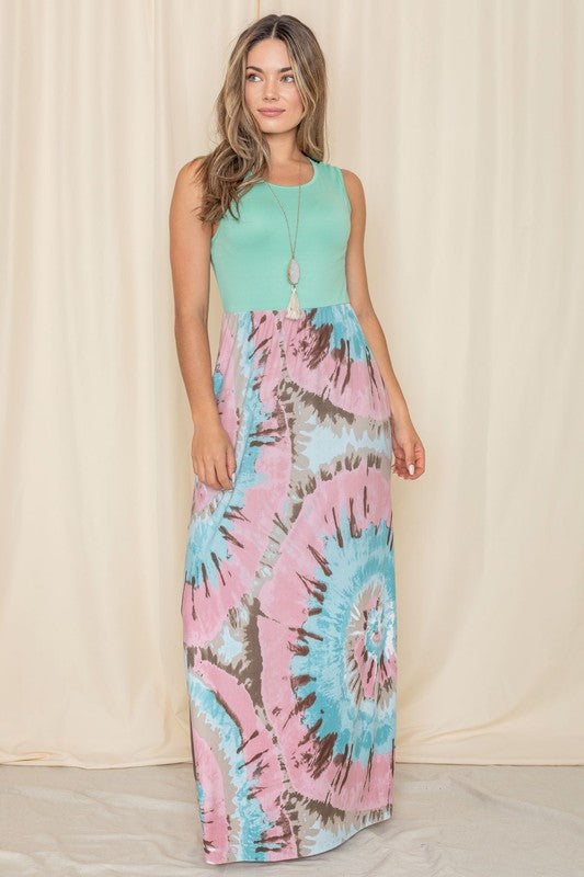 A woman is wearing the Plus Summer Sleeveless Maxi Dress, which features a mint green top paired with a skirt in a pink, blue, and brown tie-dye pattern. This American-made maxi dress, with its sleeveless design, stands out against the neutral background.