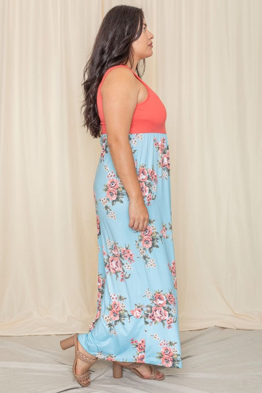 A woman elegantly stands in front of a draped curtain, wearing the Plus Sleeveless Floral Maxi Dress, which features a vibrant red top paired with a flowing blue skirt.