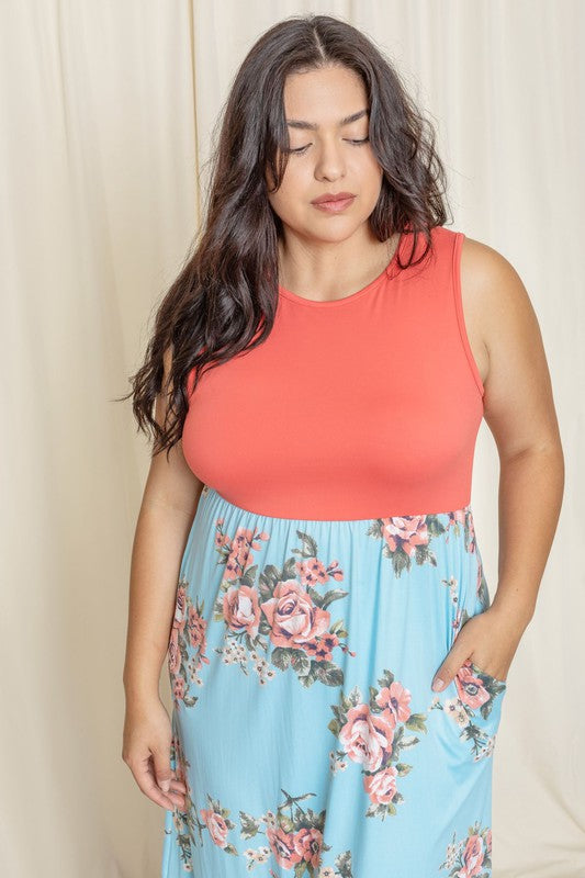 A woman elegantly stands in front of a draped curtain, wearing the Plus Sleeveless Floral Maxi Dress, which features a vibrant red top paired with a flowing blue skirt.