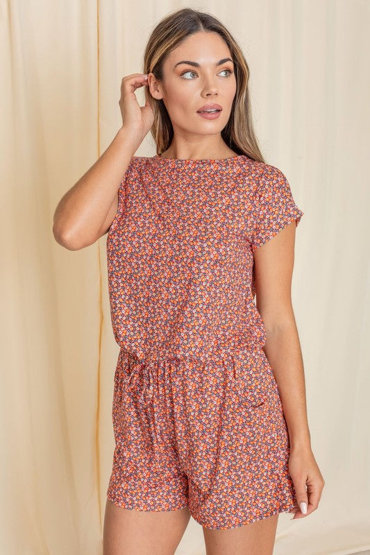 A person wearing a Short Sleeve Floral Print Romper and white sneakers stands in front of a beige curtain, showcasing an effortlessly chic style.