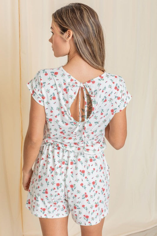 A person wearing a Short Sleeve Floral Print Romper and white sneakers stands in front of a beige curtain, showcasing an effortlessly chic style.