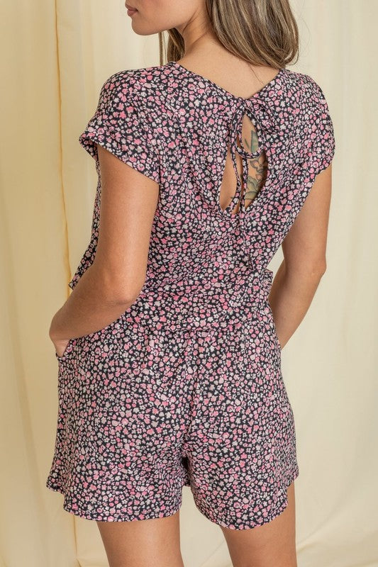 A person wearing a Short Sleeve Floral Print Romper and white sneakers stands in front of a beige curtain, showcasing an effortlessly chic style.