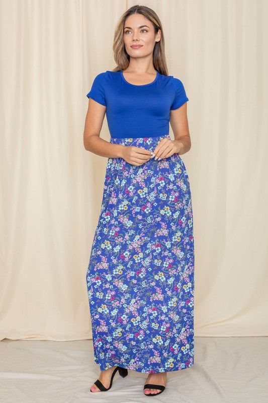 A person stands gracefully against a beige curtain backdrop, wearing a short sleeve floral maxi dress.