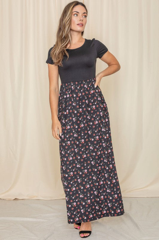 A person stands gracefully against a beige curtain backdrop, wearing a short sleeve floral maxi dress.