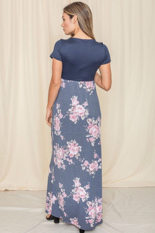 A person stands gracefully against a beige curtain backdrop, wearing a short sleeve floral maxi dress.