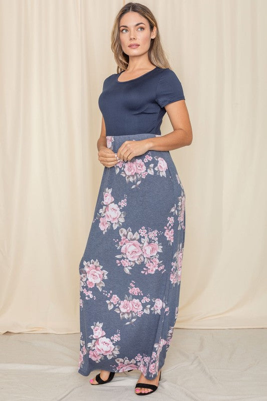 A person stands gracefully against a beige curtain backdrop, wearing a short sleeve floral maxi dress.