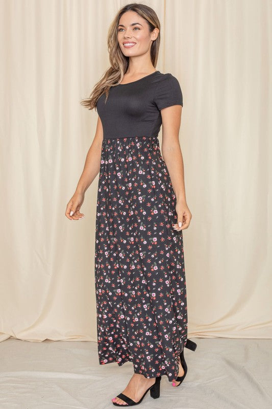 A person stands gracefully against a beige curtain backdrop, wearing a short sleeve floral maxi dress.