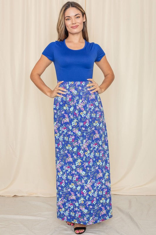 A person stands gracefully against a beige curtain backdrop, wearing a short sleeve floral maxi dress.