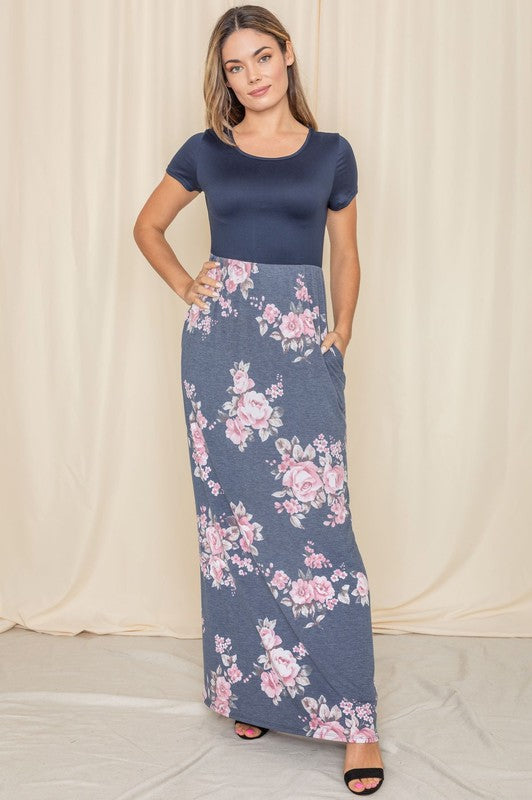 A person stands gracefully against a beige curtain backdrop, wearing a short sleeve floral maxi dress.