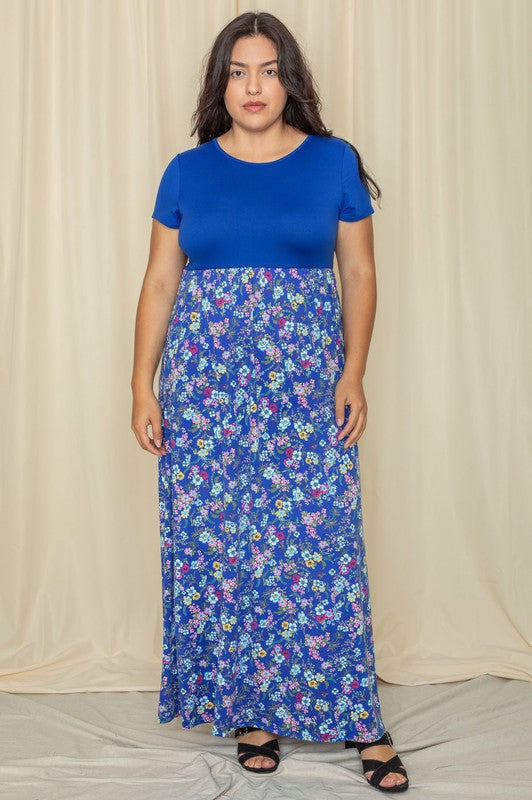 A person wearing a Plus Short Sleeve Floral Maxi Dress, featuring a blue floral pattern, smiles with hands in pockets while standing in front of a beige curtain.