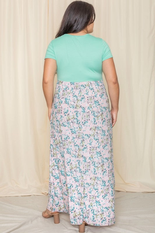 A person wearing the Plus Short Sleeve Floral Maxi Dress, featuring a gray top and flowing floral design, stands gracefully in front of a beige curtain.