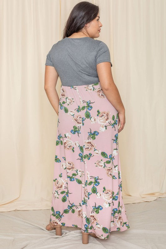 A person wearing the Plus Short Sleeve Floral Maxi Dress, featuring a gray top and flowing floral design, stands gracefully in front of a beige curtain.