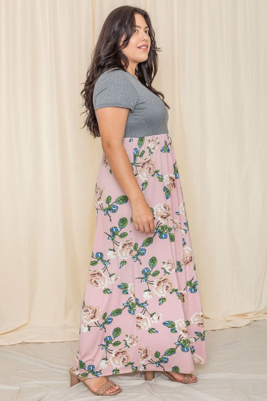 A person wearing the Plus Short Sleeve Floral Maxi Dress, featuring a gray top and flowing floral design, stands gracefully in front of a beige curtain.