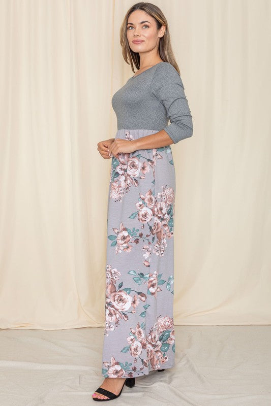 A woman stands against a beige backdrop wearing the elegant Quarter Sleeve Floral Maxi Dress. It showcases a stunning gray top with quarter sleeves and a beautifully patterned floral skirt, seamlessly blending elegance and style. Made in the United States, this dress is truly captivating.