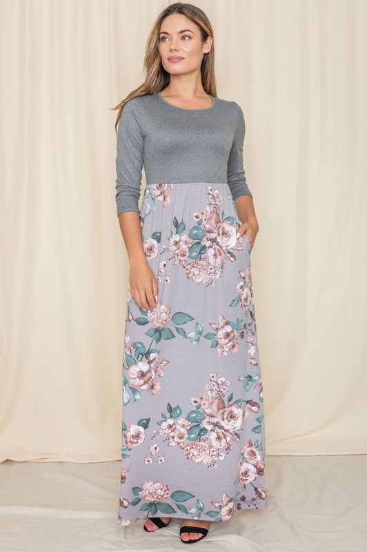 A woman stands against a beige backdrop wearing the elegant Quarter Sleeve Floral Maxi Dress. It showcases a stunning gray top with quarter sleeves and a beautifully patterned floral skirt, seamlessly blending elegance and style. Made in the United States, this dress is truly captivating.