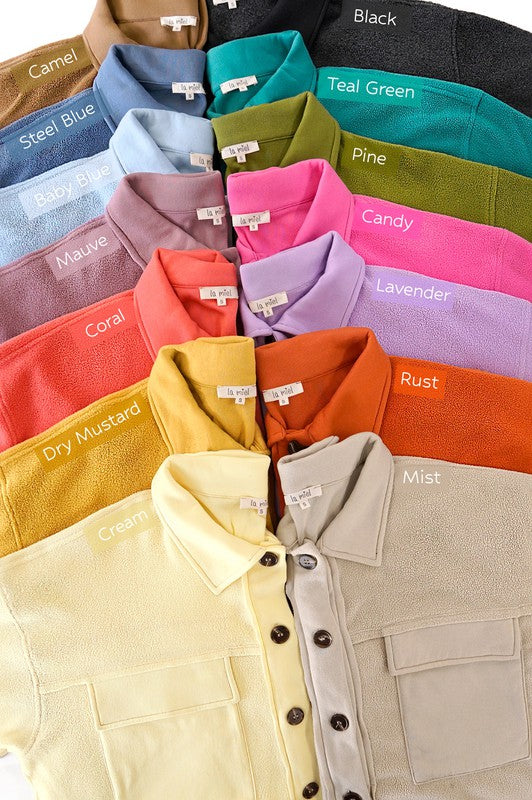 A woman in an oversized, lightweight Matilda Jacket in beige with a button-up front and an orange hat sits against a green background with her eyes closed. She is sporting a bright orange top and white shorts, embracing a casual style.