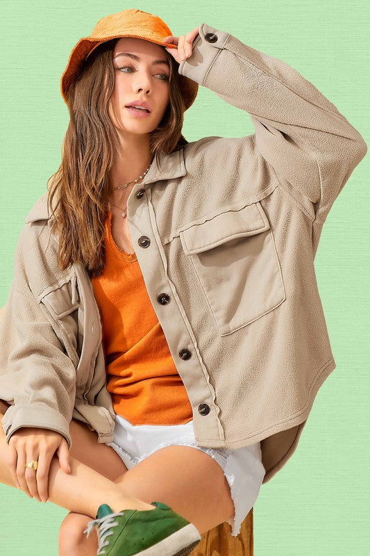 A woman in an oversized, lightweight Matilda Jacket in beige with a button-up front and an orange hat sits against a green background with her eyes closed. She is sporting a bright orange top and white shorts, embracing a casual style.