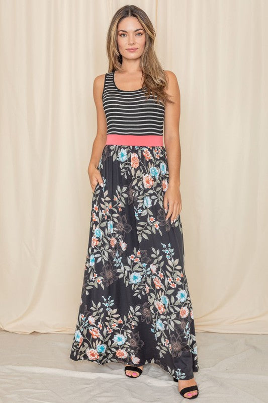 A woman wearing the Sleeveless Floral Band Maxi Dress, featuring a striped top, floral skirt, and coral waistband, stands against a beige backdrop.