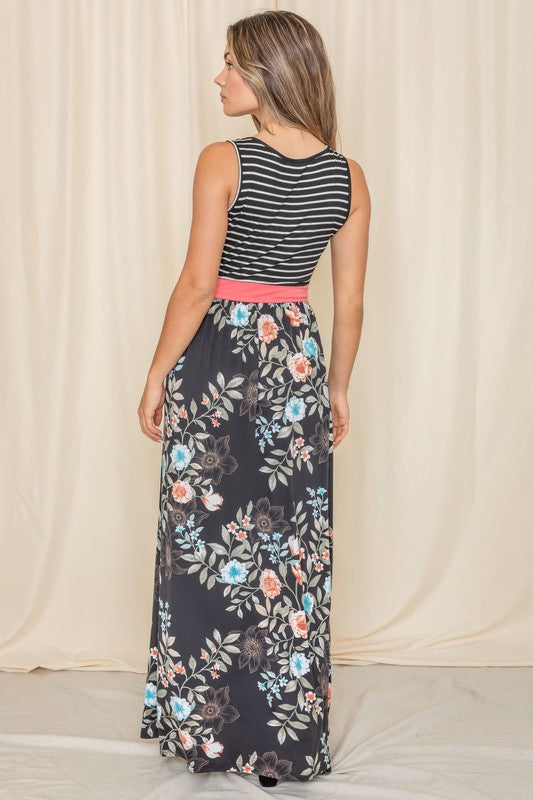 A woman wearing the Sleeveless Floral Band Maxi Dress, featuring a striped top, floral skirt, and coral waistband, stands against a beige backdrop.