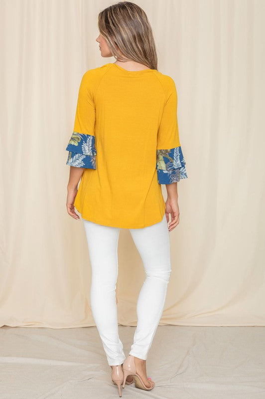 A woman wearing a Floral Double Ruffle Tunic with yellow fabric and floral-patterned sleeve cuffs stands against a beige curtain. Crafted with care in the United States, her style embodies a perfect blend of elegance and comfort.