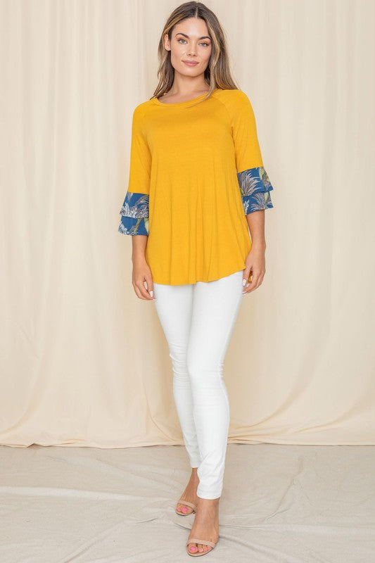 A woman wearing a Floral Double Ruffle Tunic with yellow fabric and floral-patterned sleeve cuffs stands against a beige curtain. Crafted with care in the United States, her style embodies a perfect blend of elegance and comfort.