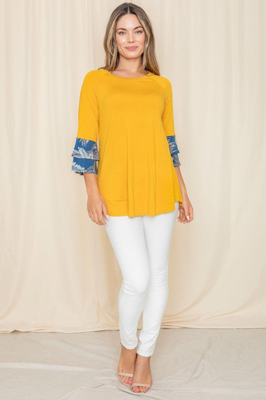 A woman wearing a Floral Double Ruffle Tunic with yellow fabric and floral-patterned sleeve cuffs stands against a beige curtain. Crafted with care in the United States, her style embodies a perfect blend of elegance and comfort.