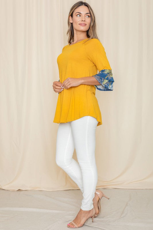 A woman wearing a Floral Double Ruffle Tunic with yellow fabric and floral-patterned sleeve cuffs stands against a beige curtain. Crafted with care in the United States, her style embodies a perfect blend of elegance and comfort.