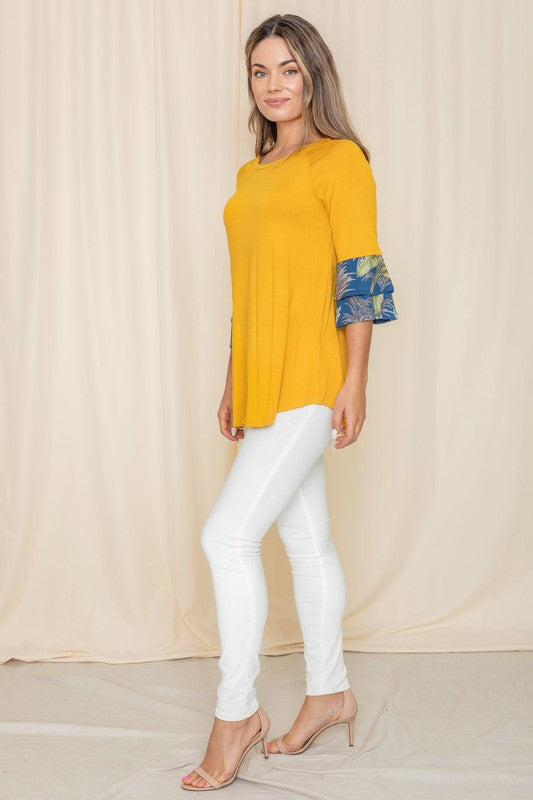 A woman wearing a Floral Double Ruffle Tunic with yellow fabric and floral-patterned sleeve cuffs stands against a beige curtain. Crafted with care in the United States, her style embodies a perfect blend of elegance and comfort.