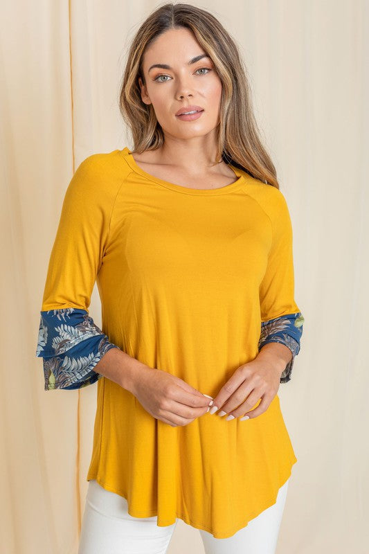 A woman wearing a Floral Double Ruffle Tunic with yellow fabric and floral-patterned sleeve cuffs stands against a beige curtain. Crafted with care in the United States, her style embodies a perfect blend of elegance and comfort.