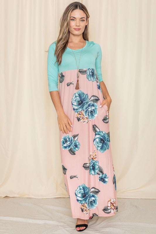 A woman elegantly poses in a High Waist Floral Maxi Dress, featuring a lovely teal hue, against a backdrop of light-colored curtains.