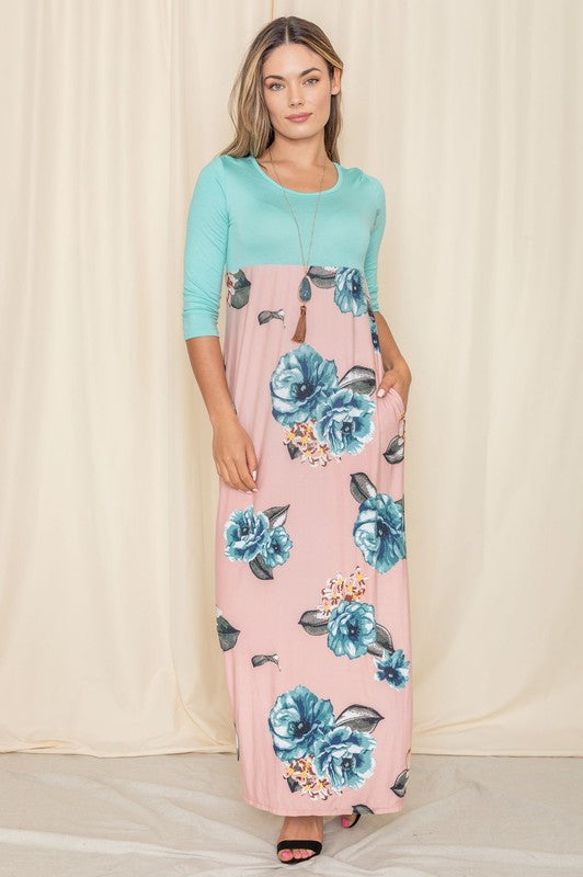 A woman elegantly poses in a High Waist Floral Maxi Dress, featuring a lovely teal hue, against a backdrop of light-colored curtains.