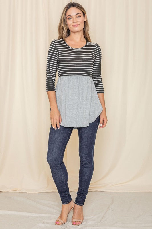 A woman elegantly stands in front of a beige curtain backdrop, wearing a stylish black striped top with a gray peplum and dark jeans, highlighting the sophistication of the Stripe Babydoll Tunic, proudly crafted in the United States.