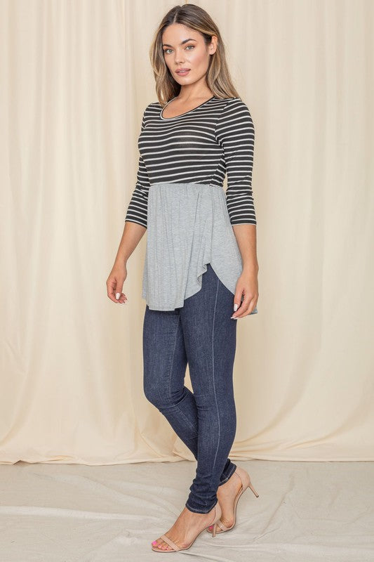 A woman wearing the Stripe Babydoll Tunic, designed with Rayon Spandex and featuring a gray flowy bottom, stands in front of a light curtain, gazing to the side.